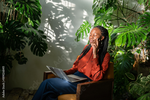Valokuva Relaxed African American woman with closed eyes dreaming and thinking while sitting with laptop in home green orangery filled with various tropical plants