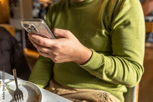 A person sits at a table in a cafe looking at their phone while food remains on their plate. They are focused on scrolling through social media content.
