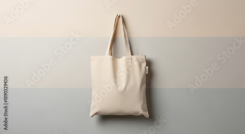 A white tote bag hangs from a strap against a wall with a beige and gray background viewed from the front