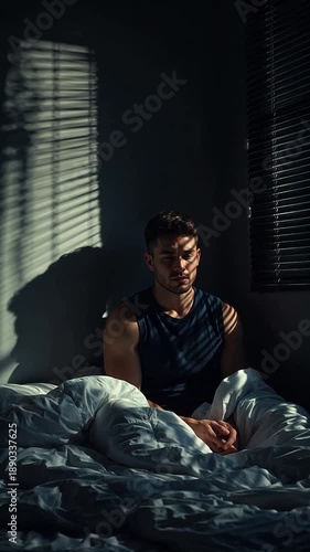 Man sitting on bed in dark room with sunlight streaming through blinds