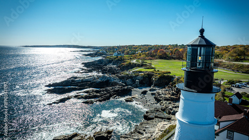 Aerial view of the iconic Portland Head Light standing tall against the rugged coastline, where the Atlantic's sapphire waves crash against dark rocks, Cape Elizabeth, Maine, United States.