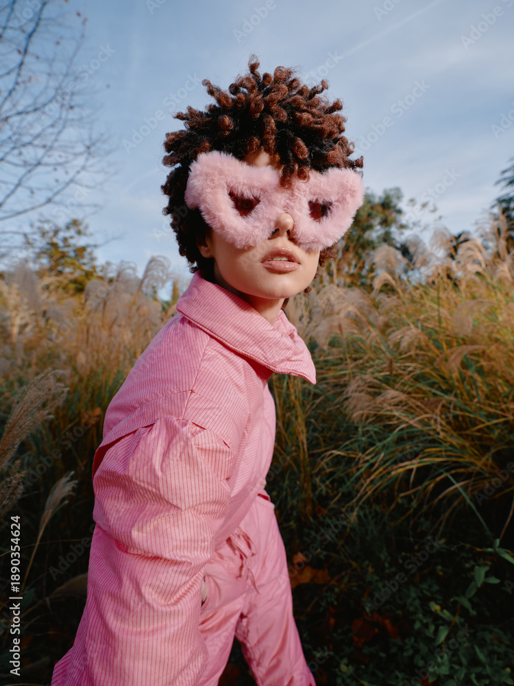 Fototapeta premium Child wearing a pink outfit and fluffy pink mask in an outdoor meadow, posing with curiosity among tall grasses and a bright sky, conveying imaginative dress up and playful fashion in nature