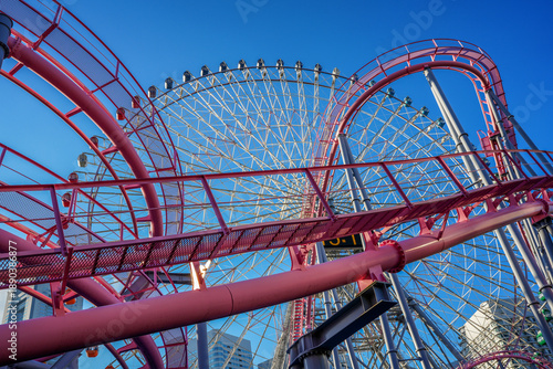 A close-up of a roller coaster and Ferris wheel at an amusement park in a tourist port city in Japan against a blue sky on a sunny day.