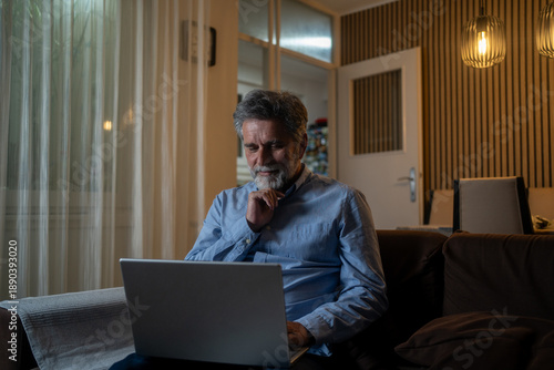 Concentrated mature bearded man sitting in home using laptop computer.