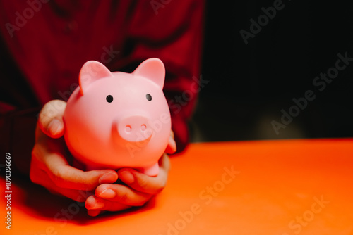 Saving concept. Pink piggy bank hands saving money finance investment protection orange table dark background person holding close up