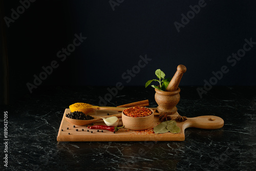 Professional still life photography showcasing assorted culinary spices and fresh herbs arranged on wooden cutting board featuring mortar and pestle on dark marble against black background