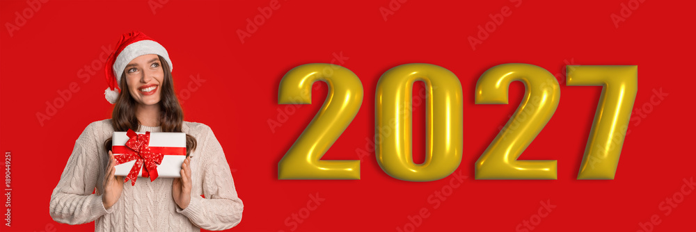 Naklejka premium A woman wearing a festive hat smiles while holding a gift in front of a bright red background with the year 2027 displayed. This scene captures a joyful moment during a holiday celebration.
