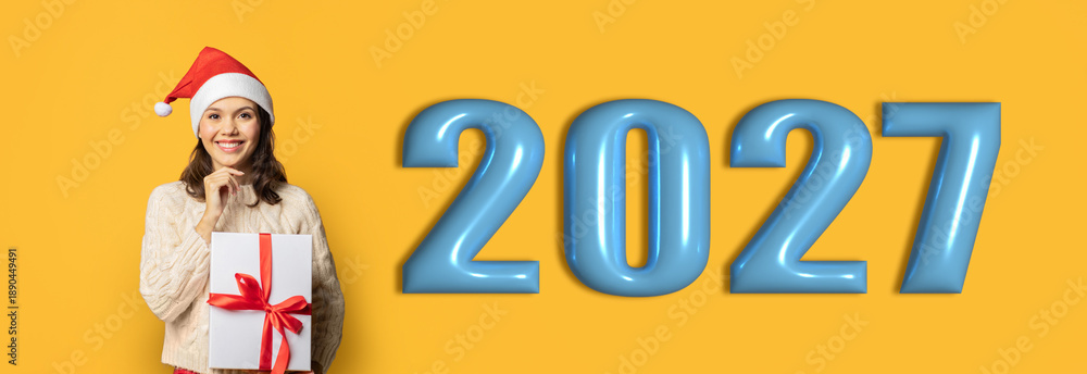 Naklejka premium A person wearing a Santa hat holds a gift wrapped with a red ribbon while smiling. The background shows a bright yellow color with large numbers representing the year 2027.