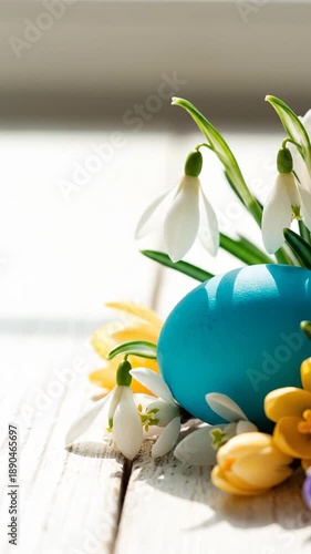 Bright blue egg placed among white flowers and yellow decorations. Sunlight shines on light wooden surface. Concept of spring, Easter, holiday celebrations