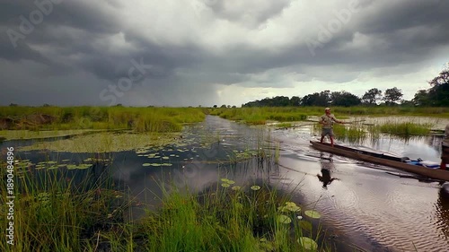 Travelers navigate the shallow waters of Okavango Delta using a canoe. They walk along the banks while observing the landscape and wildlife under dramatic skies.