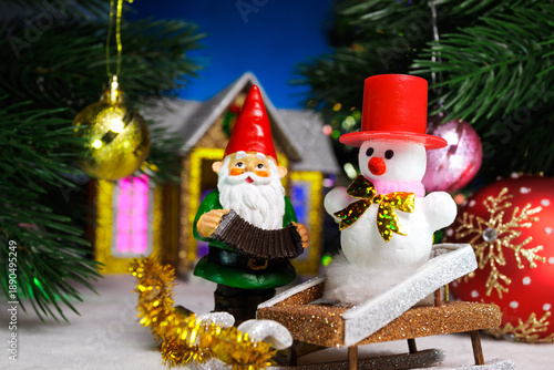 A gnome with a snowman on a sled between two balls hanging on Christmas trees against the backdrop of a Christmas house