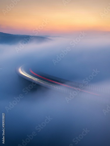 Aerial view of a bridge piercing through a dense fog blanket, light trails painting streaks across the misty canvas, Crni Kal, Koper, Slovenia.