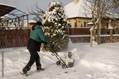 A senior man clears snow near his house. A person works with a snowplow. There is a lot of snow; winter, housing estate.