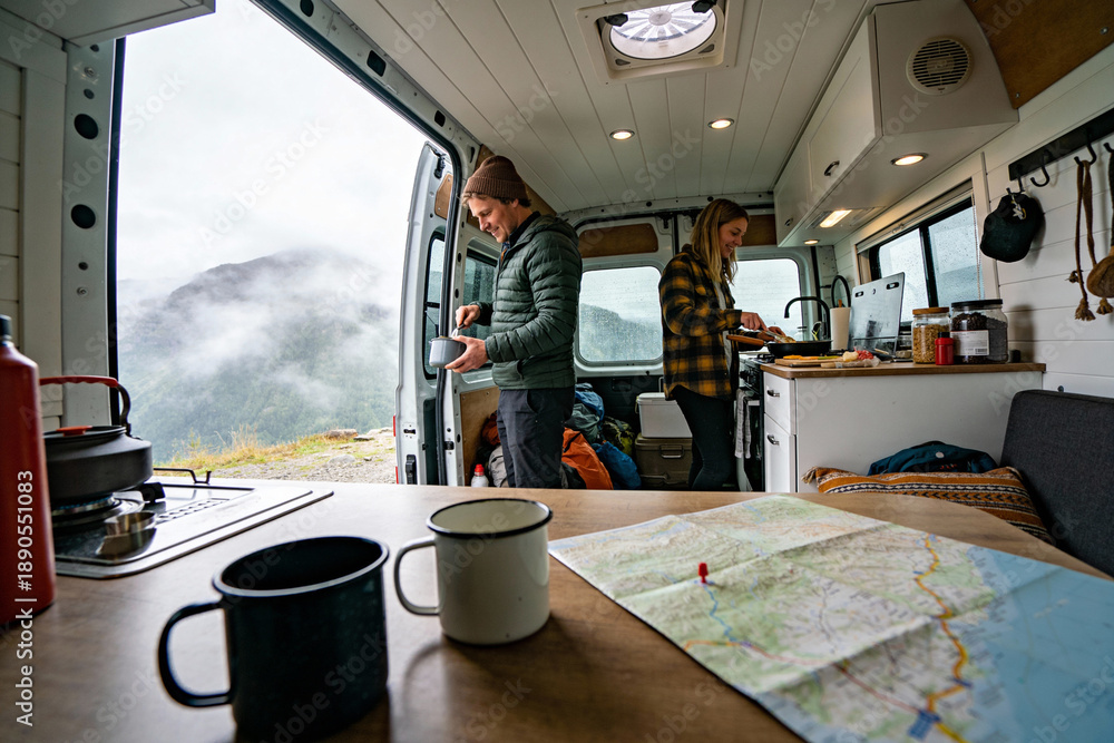 Fototapeta premium Two people prepare drinks inside a camper van with a scenic foggy mountain view outside the open door. Concept of adventure, travel, camping, and outdoor lifestyle.