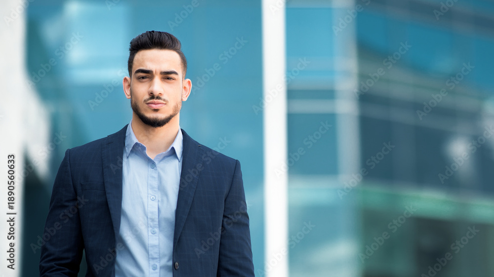 Fototapeta premium Handsome middle-eastern young businessman in suit posing next to office center, serious arabic entrepreneur having break after successful busienss meeting, standing on street, copy space, panorama