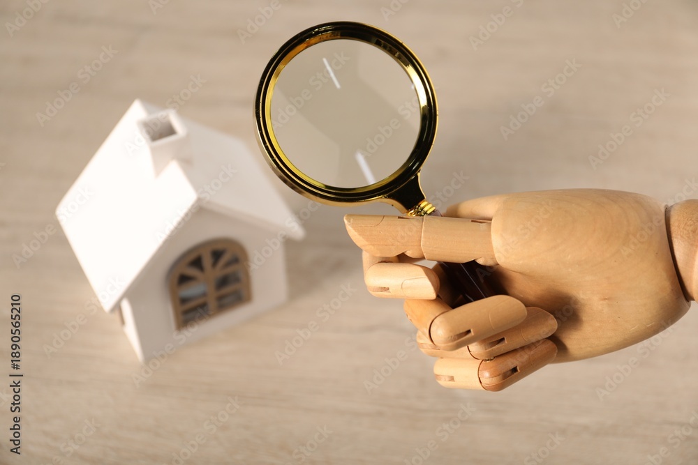 Fototapeta premium Mannequin hand with magnifying glass near house model at wooden table, above view