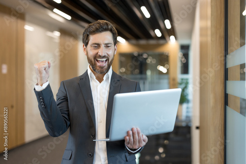 Wallpaper Mural Businessman standing in an office corridor, happily celebrating success and achieving goals while looking at his laptop, feeling triumph and professional accomplishment Torontodigital.ca
