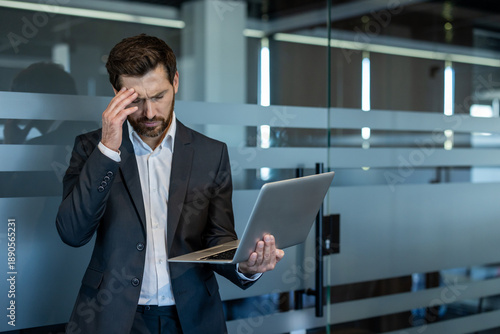 Stressed businessman in a suit suffering from a headache and work pressure while holding a laptop, standing by a frosted glass partition in a modern office environment