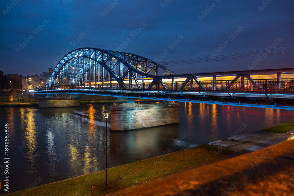 Fototapeta premium Illuminated Jozef Pilsudski Bridge in Krakow at night with train light trails and river reflection