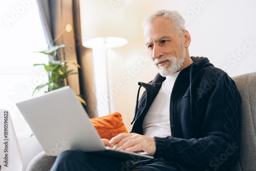 Portrait of smiling senior man sitting on comfortable sofa at home using laptop, checking mail