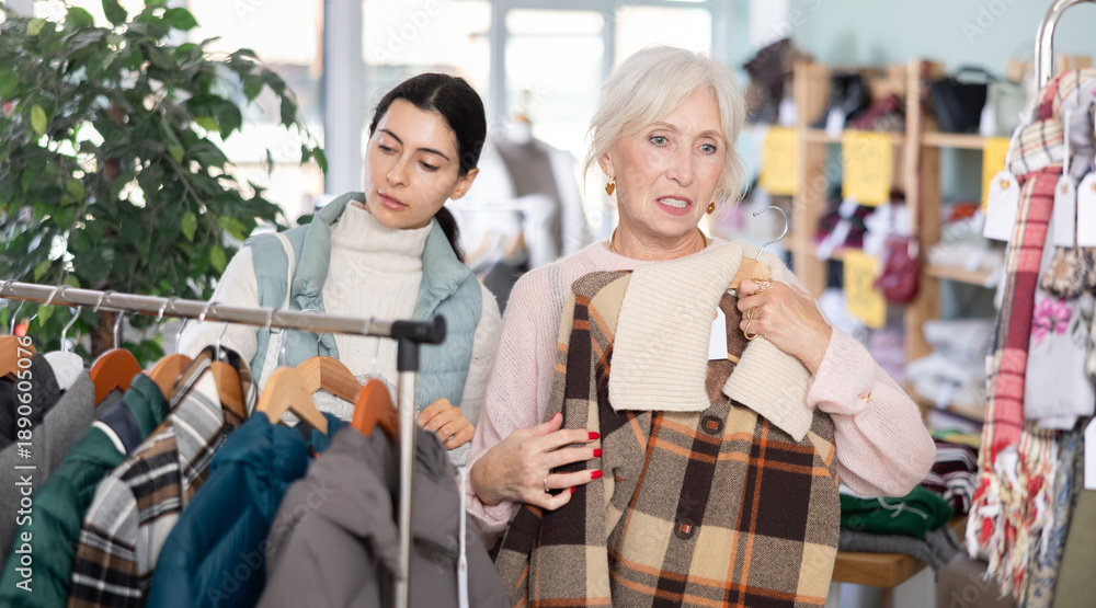 Obraz premium Two women in the store are looking at a warm coat and down jackets, discussing the quality and style among the assortment of outerwear. Mature woman and young woman buying warm clothes in a mall