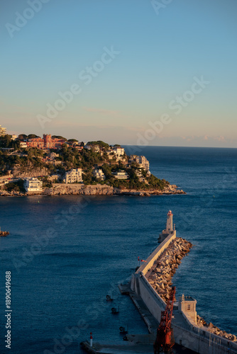 Coucher de soleil sur le port de Nice. Phare blanc dressé sur digue rocheuse, photo depuis la Coline du château avec lumière dorée, eaux méditerranéennes calmes. Quintessence Côte d'Azur. 