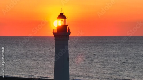AERIAL, LENS FLARE, CLOSE UP: Lighthouse tower with the setting sun shining behind the lantern room over the Mediterranean Sea. Warm golden light colors the sky, while horizon stays crisp and calm.