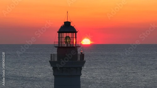 AERIAL, LENS FLARE, CLOSE UP: Sun sets behind a lighthouse, creating a vivid orange halo and sun flare over the sea. Glowing lantern and metal railings add strong graphic detail against calm horizon.