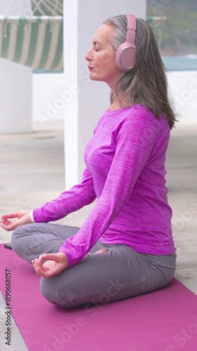 Vertical video: Sitting woman in magenta top meditating on pink mat by pool, headset, phone playing