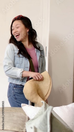 Vertical video: Hearing call, denim-clad woman pulling suitcase into entryway, holding straw hat