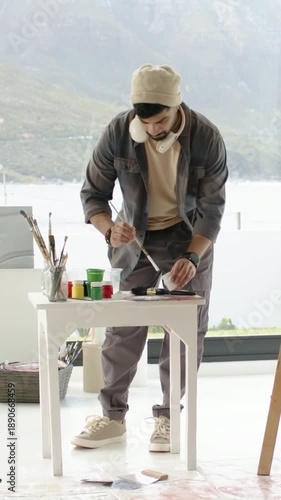Vertical video: Mixing artist preparing paints at studio window with sunlight, using cup, brushes
