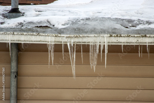 A white metal gutter in winter with an ice dam, icicles, and snow.