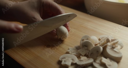Hands preparing mushrooms with a kitchen knife for homemade cooking in warm natural light, rustic kitchen atmosphere. Close-up of a chef slicing fresh white mushrooms on a wooden cutting board.