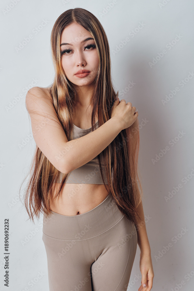 Fototapeta premium Young woman with long straight hair wearing beige sportswear posing indoors against white background. Fitness outfit, natural makeup and casual confident expression for lifestyle.