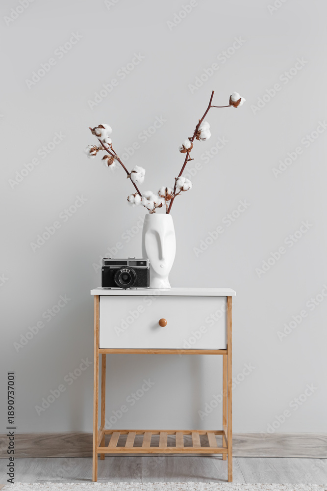 Fototapeta premium Photo camera and vase with cotton flowers on table near white wall in interior of living room