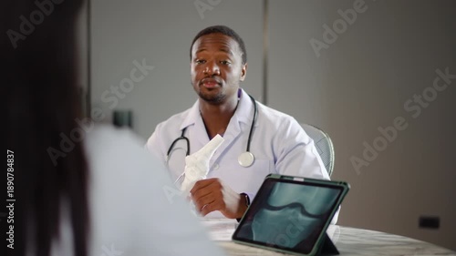 frican American orthopedic doctor consulting a female patient about knee surgery, holding an anatomical joint model and using a digital tablet with x-ray in a modern medical clinic office.