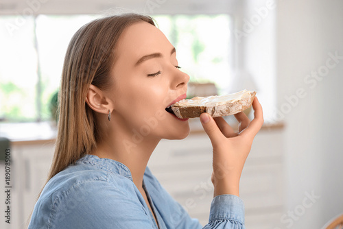 Beautiful young happy woman eating tasty toast with fresh butter in kitchen a...