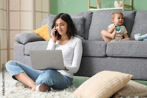 Cute baby and young mother working with laptop while talking by mobile phone ...