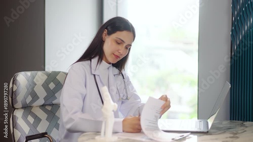 Smiling female orthopedic doctor reviews medical charts and types on a laptop in a clinic. She sits near a knee anatomy model, offering professional healthcare and happy consultation.