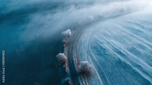 An aerial view of a snowy landscape with a winding road, the atmospheric perspective creates a sense of depth and vastness. The light and shadows give a cold, wintry atmosphere