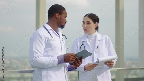 Two professional doctors, a diverse male and female team, discuss medical records on a rooftop using a tablet and clipboard, with a scenic urban cityscape background during a daytime consultation.
