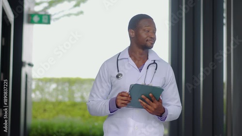 Confident Black male doctor walking in a modern hospital hallway, using a digital tablet for patient care. He smiles thoughtfully, representing healthcare innovation and medical professionalism.
