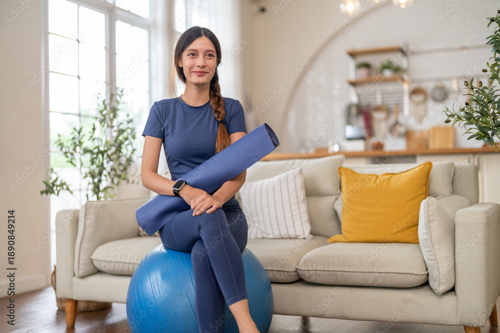 Obraz premium Happy asian woman sitting on blue exercise ball holding yoga mat ready for workout at home living room, fitness girl, healthy lifestyle, pilates swiss ball, smart watch and sport wellness concept