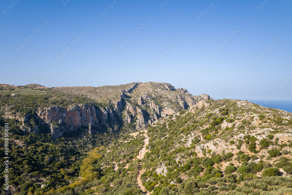 Fototapeta premium Expansive view of a rugged mountain ridge in western Crete, featuring rocky cliffs, scattered shrubs, and a winding dirt trail under a clear blue sky. The landscape is sunlit and open, evoking a sense