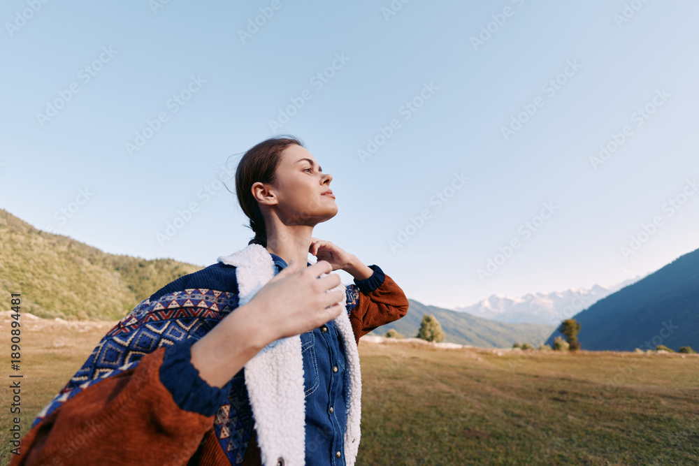 Fototapeta premium Woman standing in meadow with mountains behind, portrait of a traveler outdoors. Peaceful expression, sunlight on jacket and sweater, scenic landscape for calm relaxation.