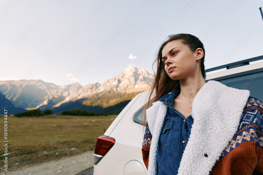 Obraz premium Woman by car in mountain landscape, travel portrait of a young woman leaning on a vehicle beside a rural road, scenic outdoor adventure with peaks, meadow and thoughtful expression