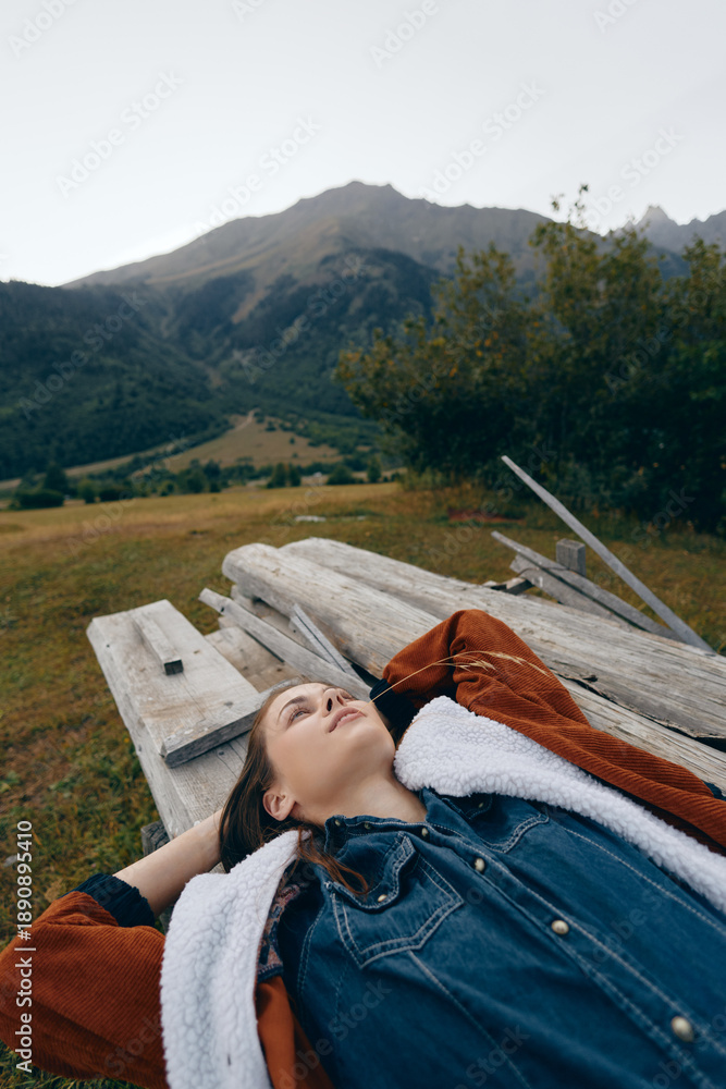 Fototapeta premium woman lying on a wooden bench in a meadow with mountains in the background, portrait of relaxation outdoors in countryside, casual denim jacket and shearling coat, peaceful scenic escape