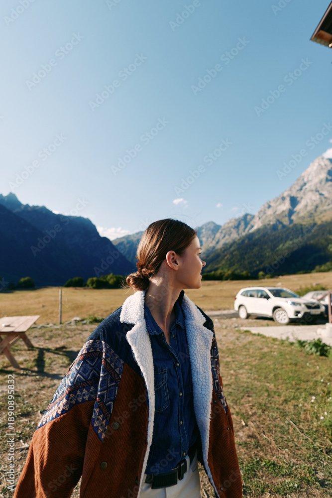 Fototapeta premium Woman in a warm jacket standing in a meadow by a white car, looking toward distant mountains during travel. Scenic outdoor portrait of nature, adventure and countryside road trip.