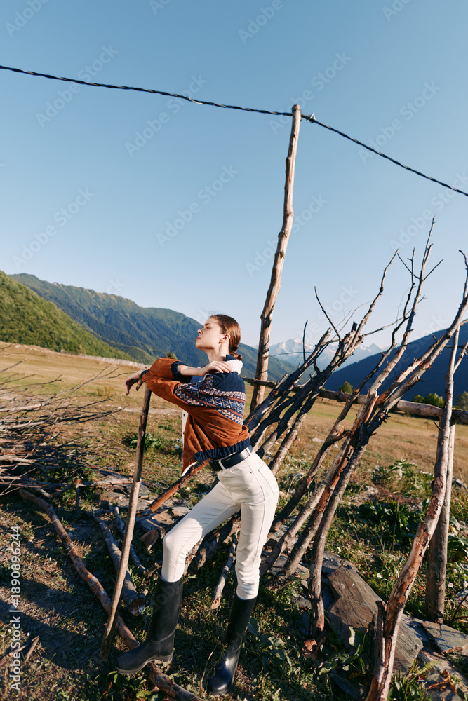Fototapeta premium Woman leans on rustic fence in countryside meadow with mountains in background, wearing sweater and boots while arranging wooden sticks for rural fence repair and outdoor work.