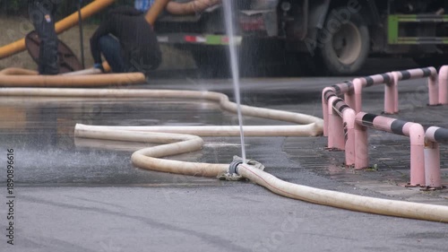 Water spraying from a damaged hose during road maintenance work, with utility workers and service trucks in background. Urban infrastructure repair maintenance, emergency water control, pipe leakage